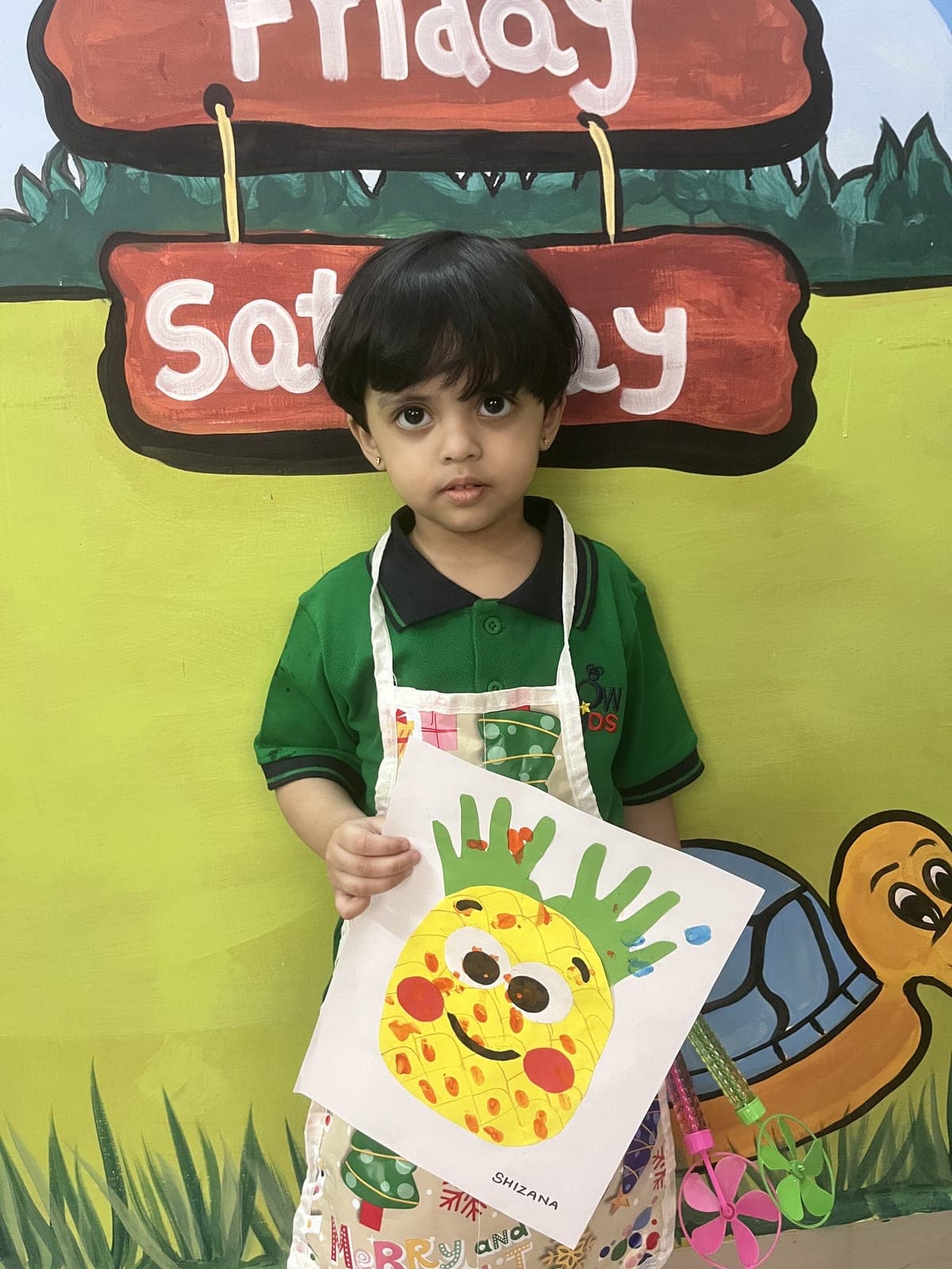 A little artist proudly holding her handprint pineapple painting in front of the colourful classroom mural