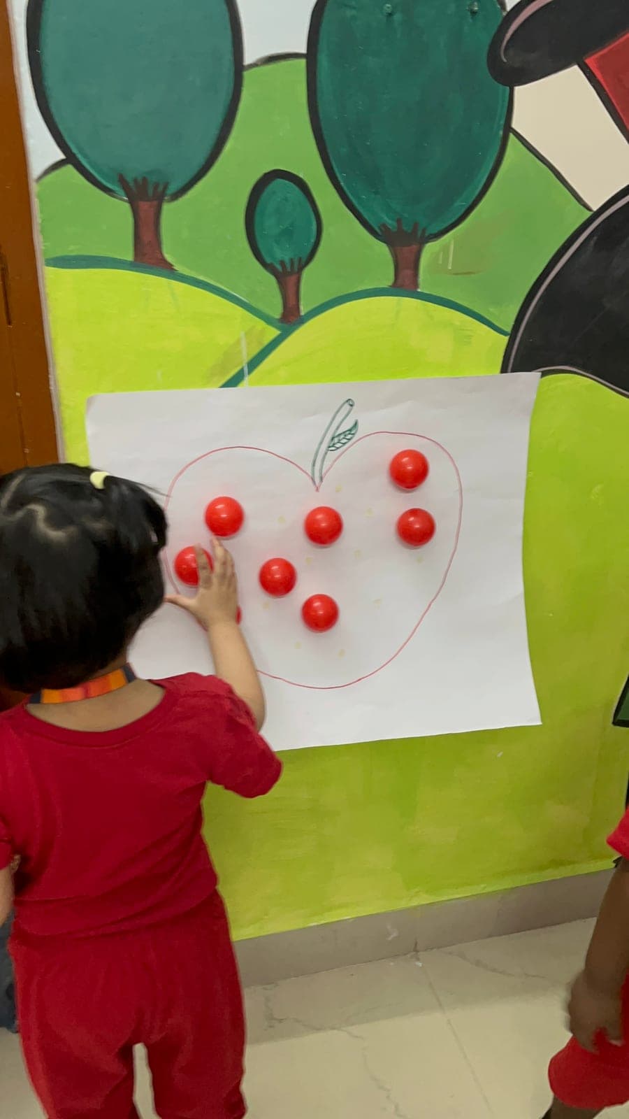 Toddler sticking red balls onto a paper apple chart to practise counting on Red Day